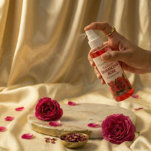 Hand holding a bottle of damascena rose water with roses and petals on a marble slab against a beige fabric background.