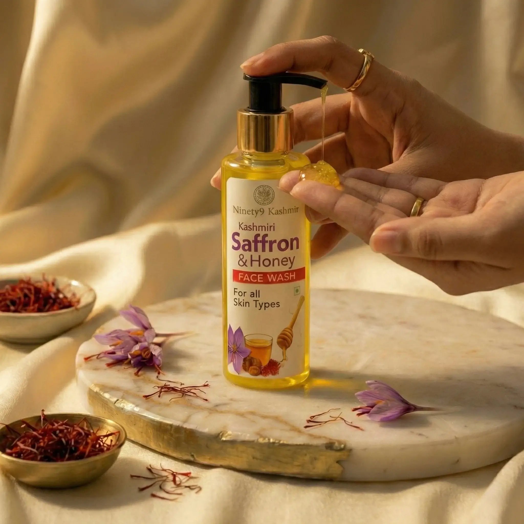 Hand holding a bottle of Kashmiri Saffron & Honey Face Wash on a marble tray with saffron and flowers.