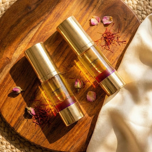Two gold Saffron Serum bottles with a purple liquid on a wooden surface with dried flowers.