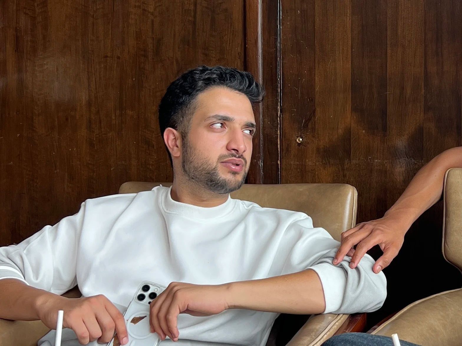 Kaunain in a white shirt sitting in a chair with another person's hand on his arm, against a wooden wall.