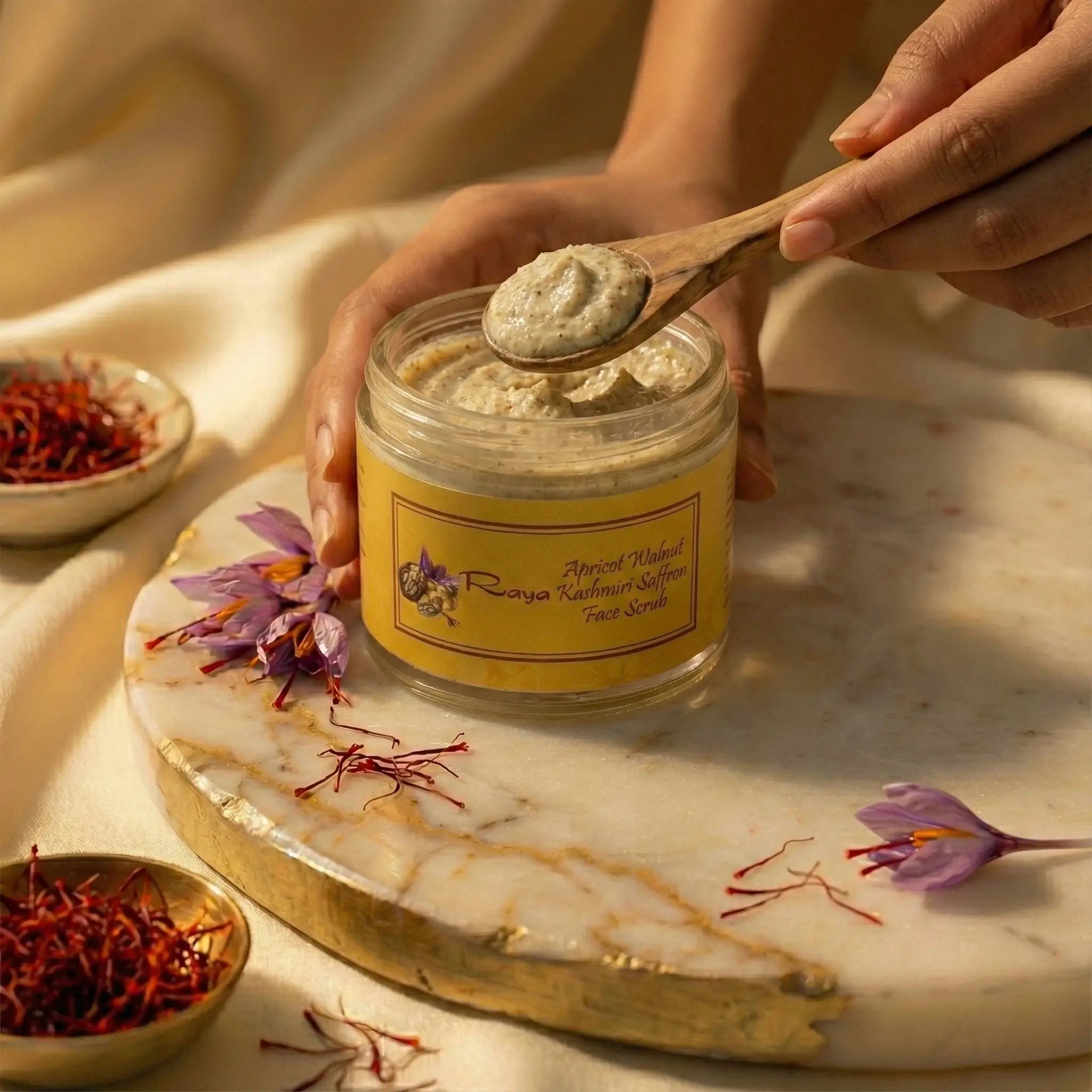 Jar of Raya face Scrub with a spoonful being taken, surrounded by saffron and flowers on a marble surface.